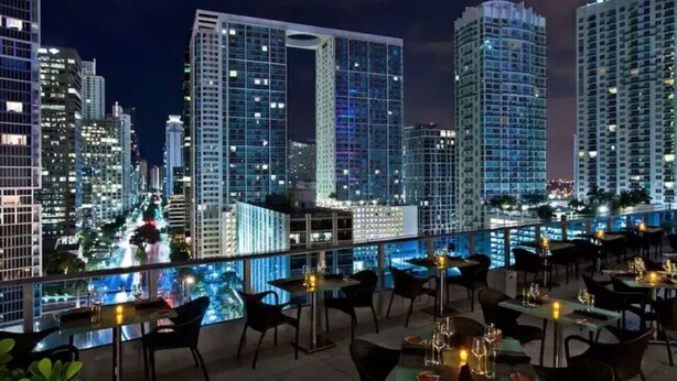 A rooftop dining area with tables and chairs, overlooking a cityscape with tall buildings and a river. - Amazing 1 Bed in Brickell key W/ FREE PKG (Miami)
