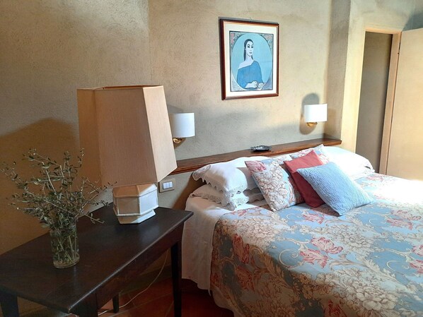 A hotel room with a bed, bedside lamps, a nightstand, a vase with dried plants, and a framed picture on the wall.