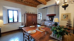 Une salle à manger avec une table en bois, des chaises et un lustre. On aperçoit une fenêtre avec des stores, un buffet et une cheminée au fond.