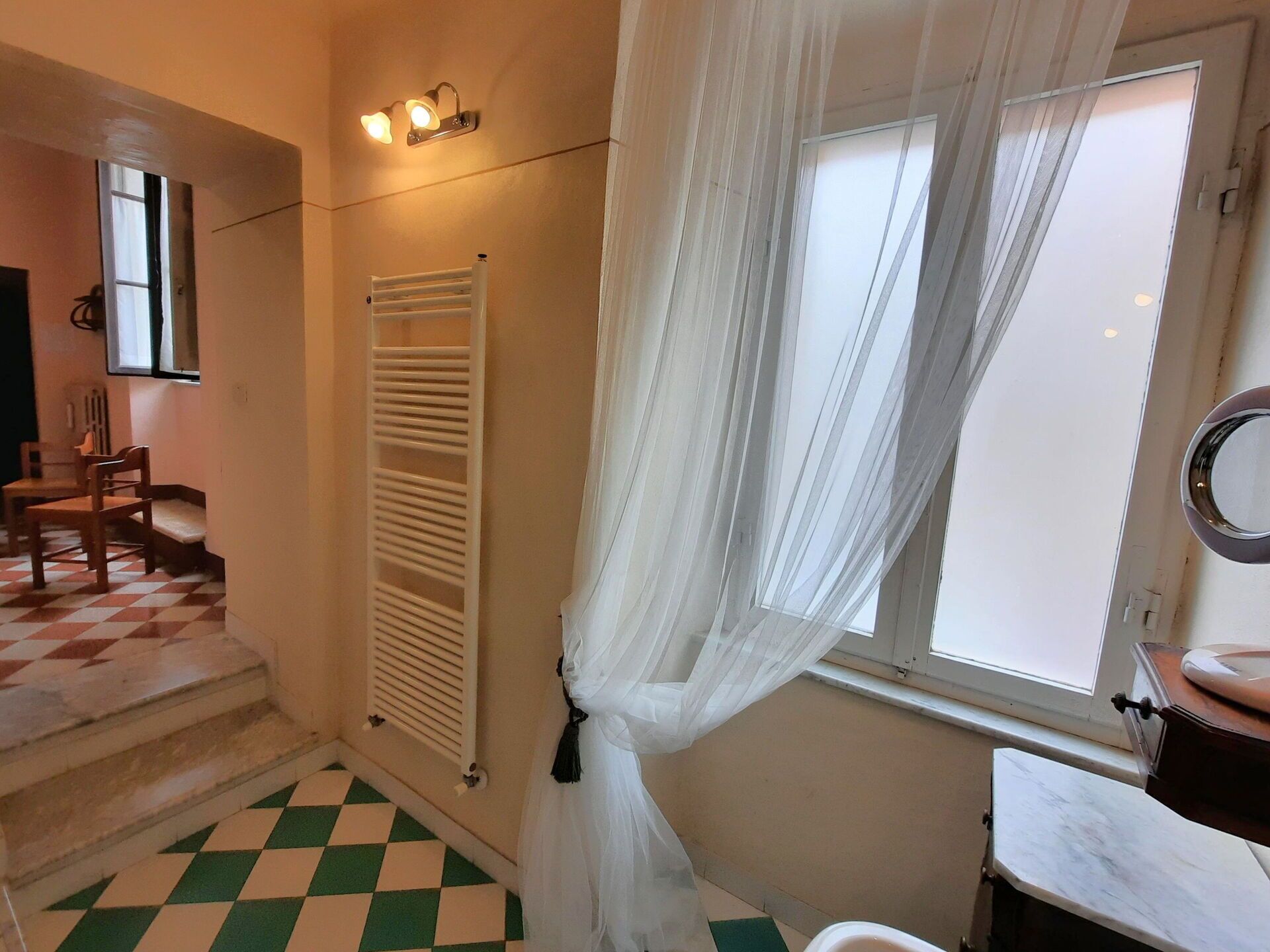 Ein Badezimmer mit Schachbrettboden, einem Heizkörper, einem Fenster mit durchscheinenden Vorhängen und einem Waschbecken mit Spiegel.