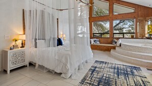 Een slaapkamer met een hemelbed, een nachtkastje met een lamp en uitzicht op de omgeving door grote ramen.