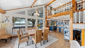 Een ruime eethoek met een grote houten tafel en rieten stoelen, een keuken met blauwe kastjes, en een balkon met uitzicht op het landschap.