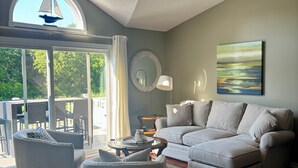 Un salon spacieux avec un grand canapé d’angle, une table basse ronde et une table d’appoint avec un plateau en verre. Une grande fenêtre offre une vue sur le mobilier d’extérieur et la verdure.
