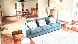 A living room with a blue sofa, white cushions, a white rug, a wooden stool, and a dining area with wicker chairs and a wooden table. - Charming house 100 mt from the famous "La Torche" beach

 (Plomeur)