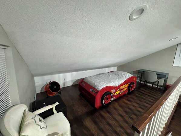 A loft bedroom with a sloped ceiling, a red toy car bed, a desk with a chair, and a white chair with a decorative pillow.