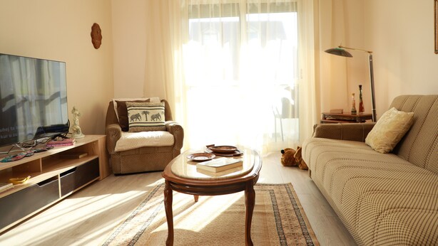 Ein Wohnzimmer mit einer Couch, einem Sessel, einem Couchtisch, einem Fernseher und einem großen Fenster mit Vorhängen.