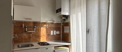 A kitchen with a gas stove, sink, and a refrigerator. There are cabinets above the counter and a window with curtains.