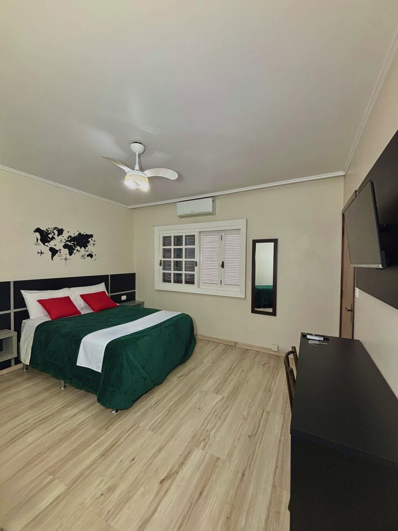A bedroom with a bed, a desk, a TV, a ceiling fan, a window with shutters, and a world map on the wall.
