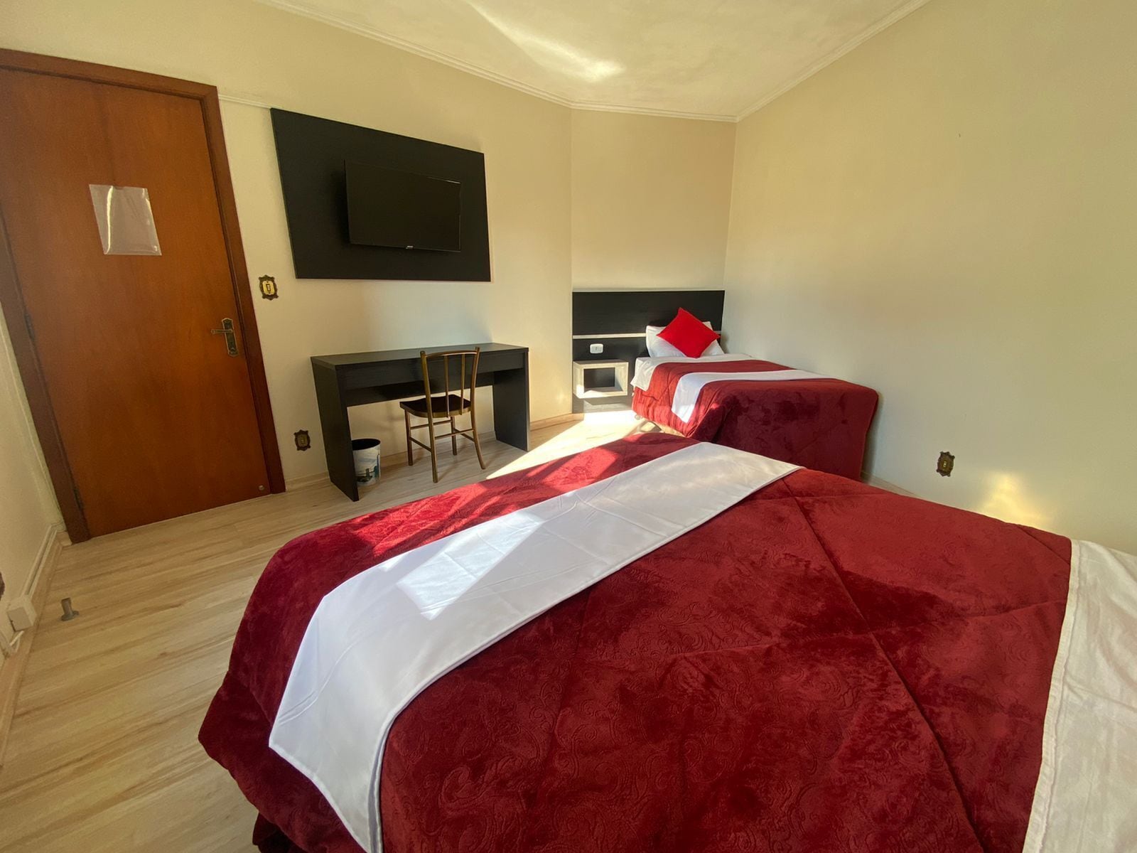 A hotel room with two beds, a desk, a chair, a TV mounted on the wall, and a wooden door.