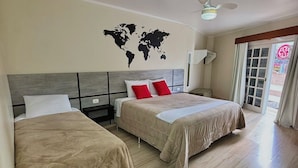 Quarto de hotel com duas camas, um mapa-múndi na parede, um ventilador de teto e vista para o exterior através de uma janela.