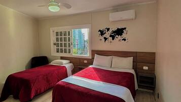 A hotel room with two beds, a ceiling fan, an air conditioning unit, a window with a view of buildings, and a world map on the wall.