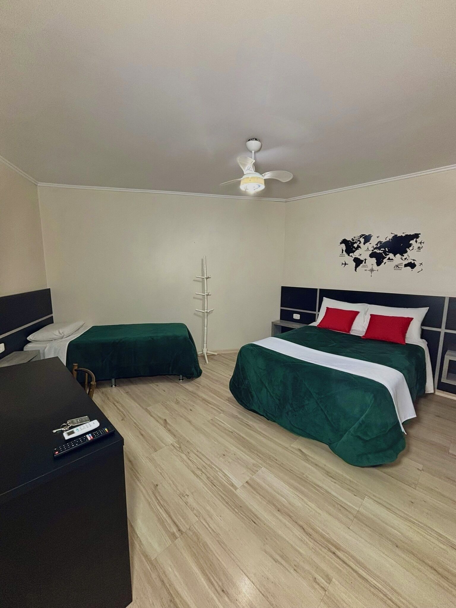 Ein Hotelzimmer mit zwei Betten, einem Schreibtisch, einem Stuhl, einem Deckenventilator und einer Weltkarte an der Wand.
