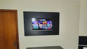 Um quarto com porta de madeira, uma escrivaninha preta com cadeira, uma televisão na parede exibindo um canal de comédia e uma pequena lixeira.