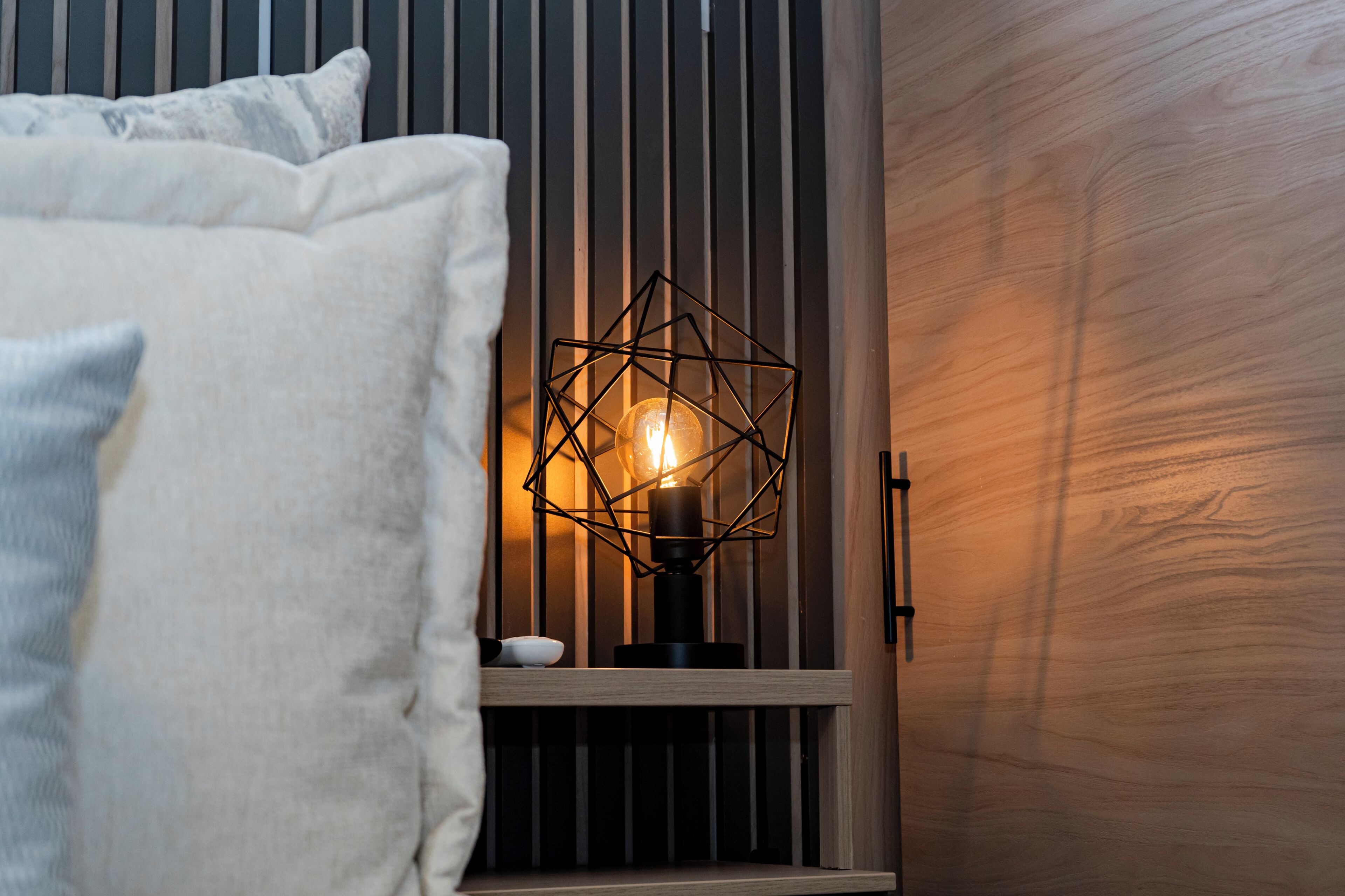 A bed with pillows, a bedside table with a geometric light fixture, and wooden paneling.