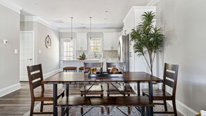 A dining area with a wooden table and chairs, a plant, and a kitchen in the background. - Pinehurst Grand Slam (West End)