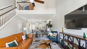 A spacious living room with a high ceiling, a staircase, a dining area, and a large TV mounted on the wall. - Pinehurst Grand Slam (West End)