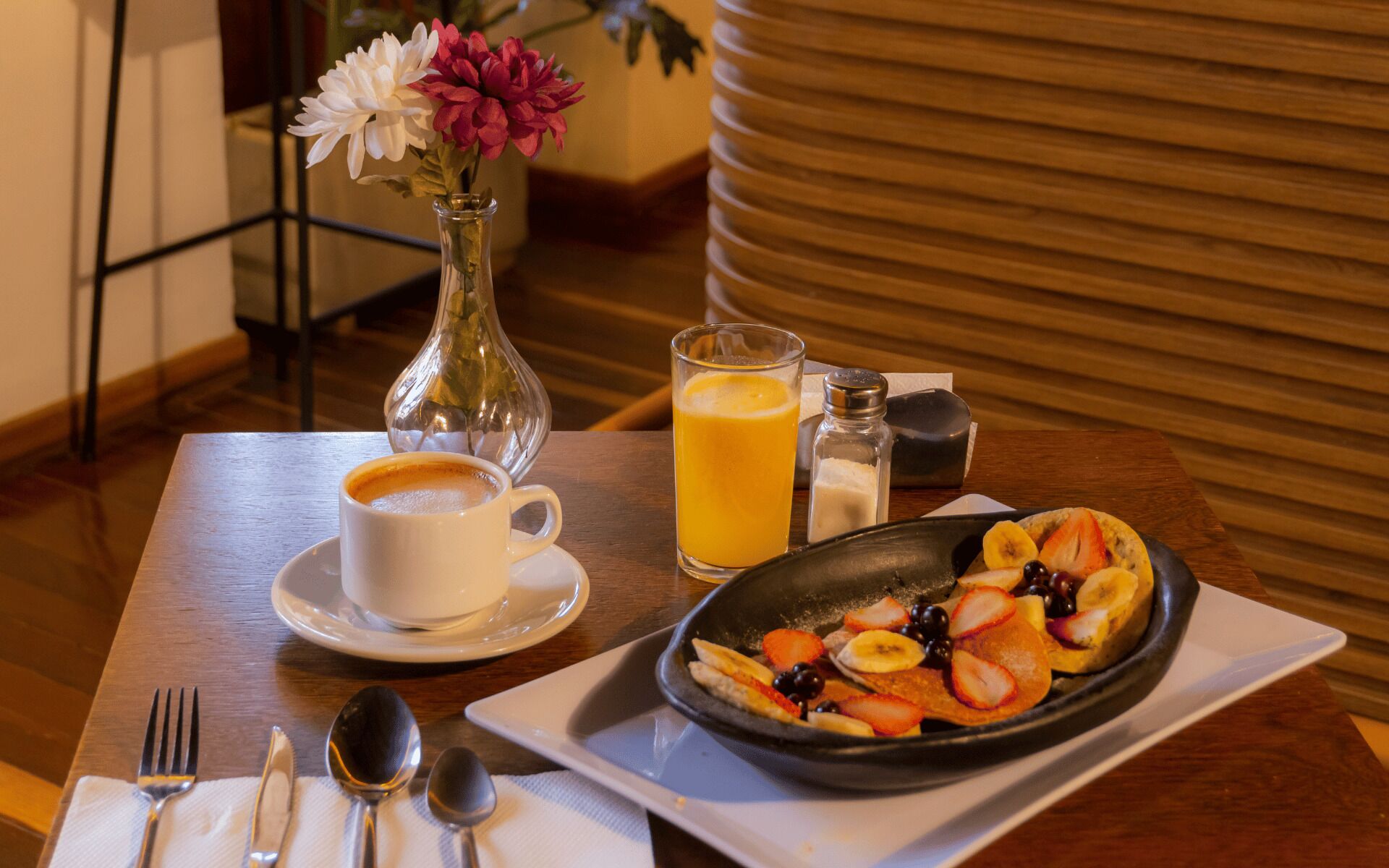 A breakfast setting with a coffee cup, orange juice, a fruit plate, a vase with flowers, and a salt and pepper shaker on a wooden table.