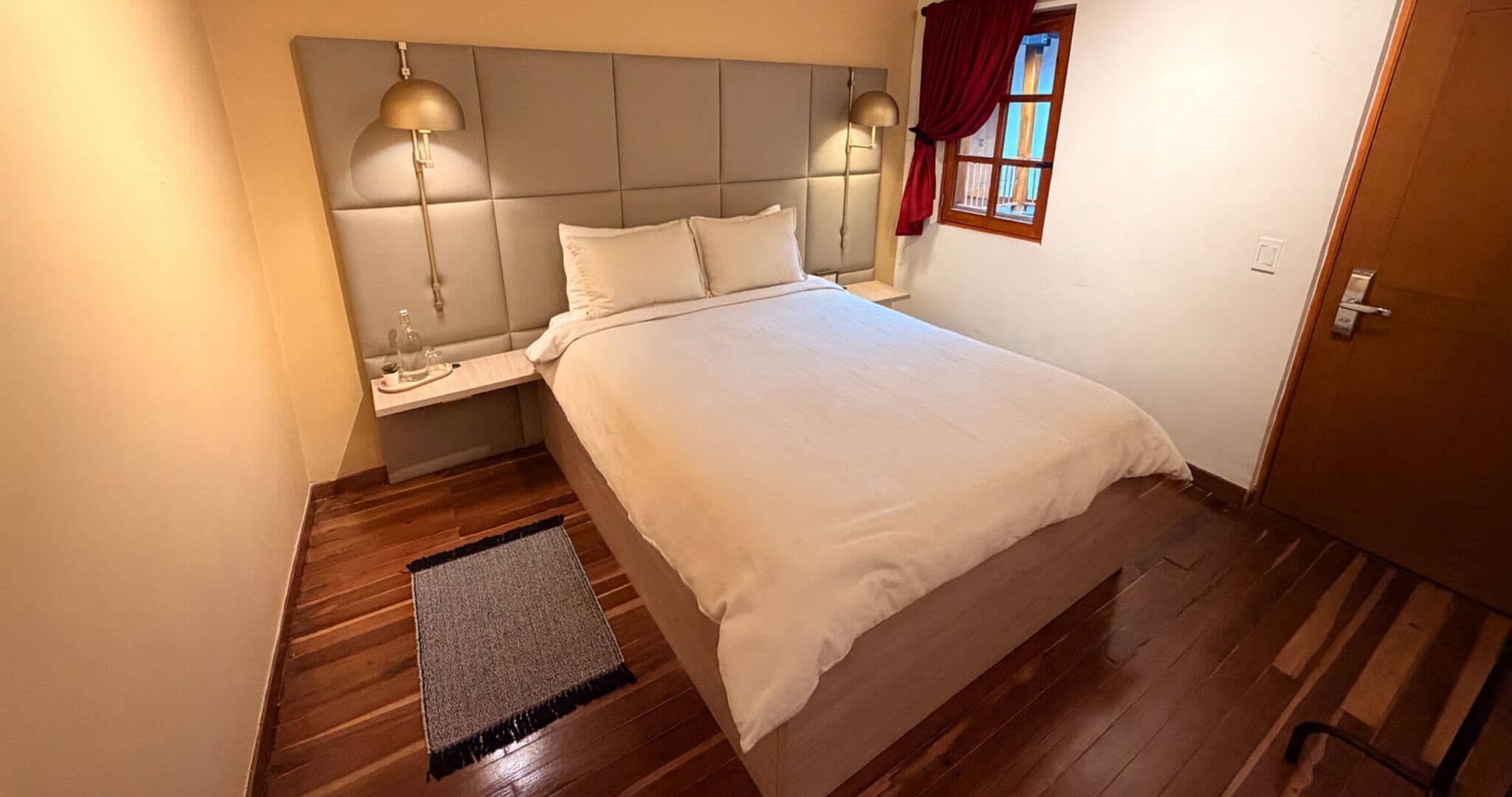 A bedroom with a bed, bedside lamps, a nightstand, a window with curtains, and a wooden floor.