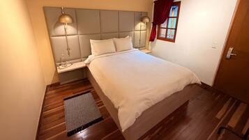 A bedroom with a bed, bedside lamps, a nightstand, a window with curtains, and a wooden floor.
