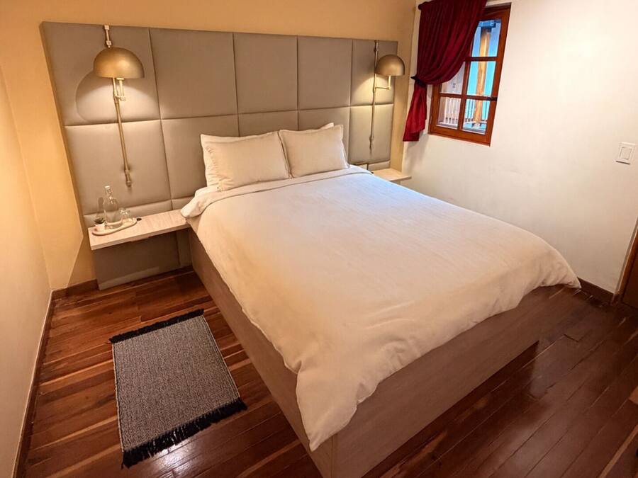 A bedroom with a bed, bedside lamps, a nightstand, a window with curtains, and a wooden floor.