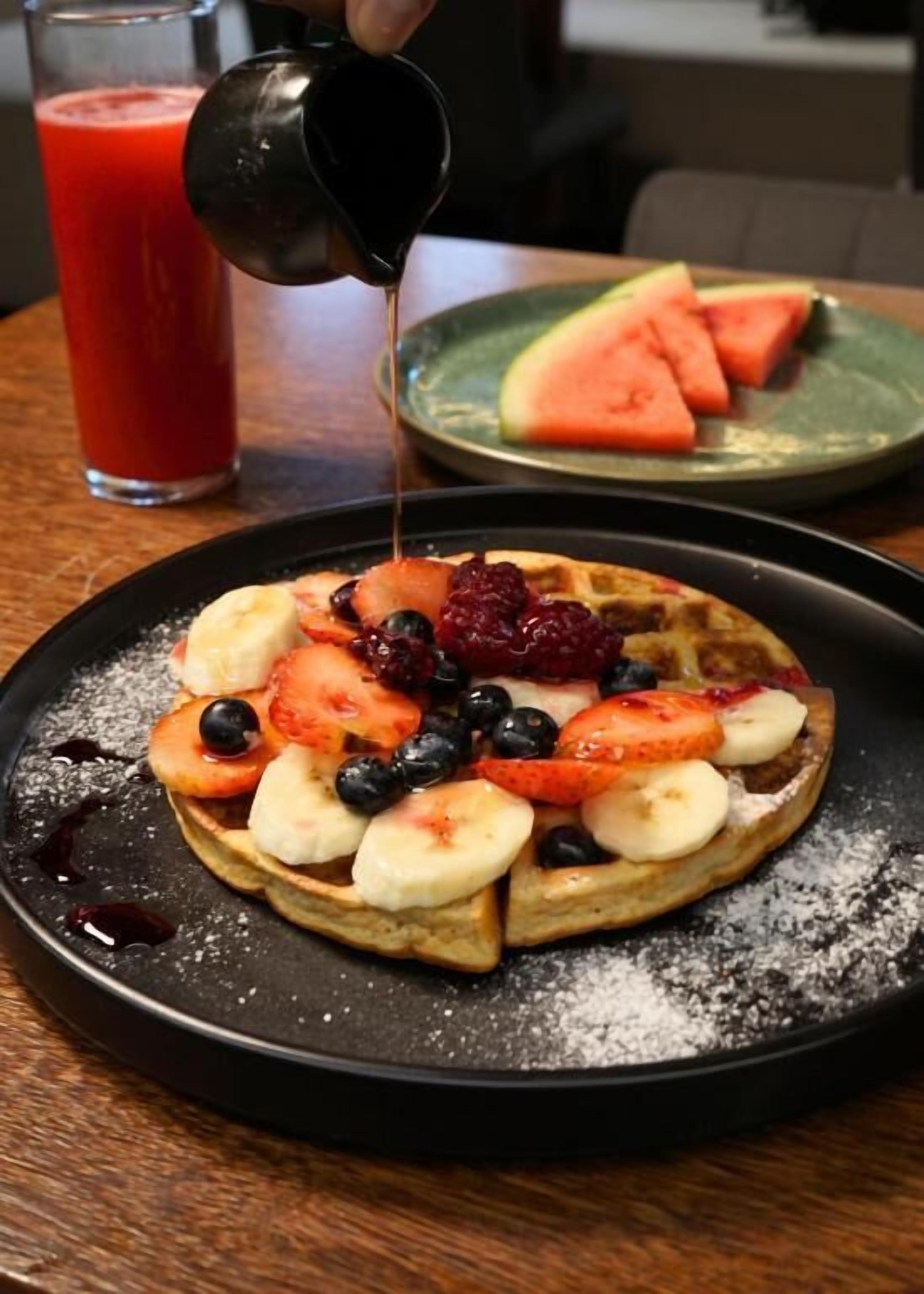 Ein Frühstücksteller mit Waffeln, belegt mit Obst und Sirup, ein Glas roter Saft und ein Teller mit Wassermelonenstücken.