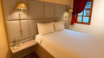A hotel room with a large bed, two bedside lamps, a small table with a bottle and glasses, and a window with red curtains.