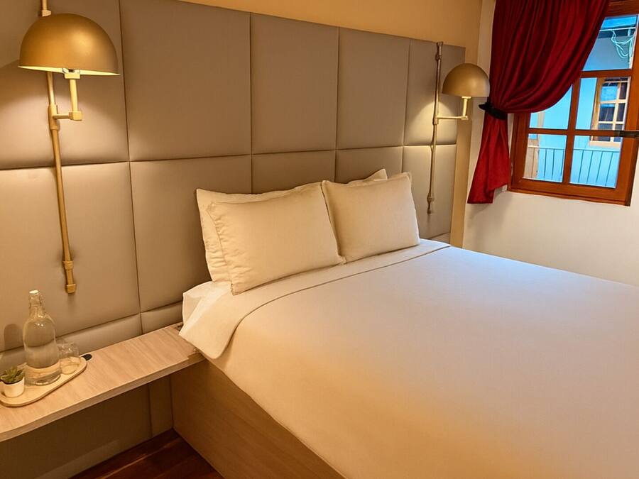A hotel room with a large bed, two bedside lamps, a small table with a bottle and glasses, and a window with red curtains.