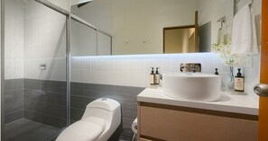 Ein modernes Badezimmer mit Duschkabine aus Glas, Toilette, Waschbecken mit Spiegel und Pflegeprodukten auf der Ablagefläche.