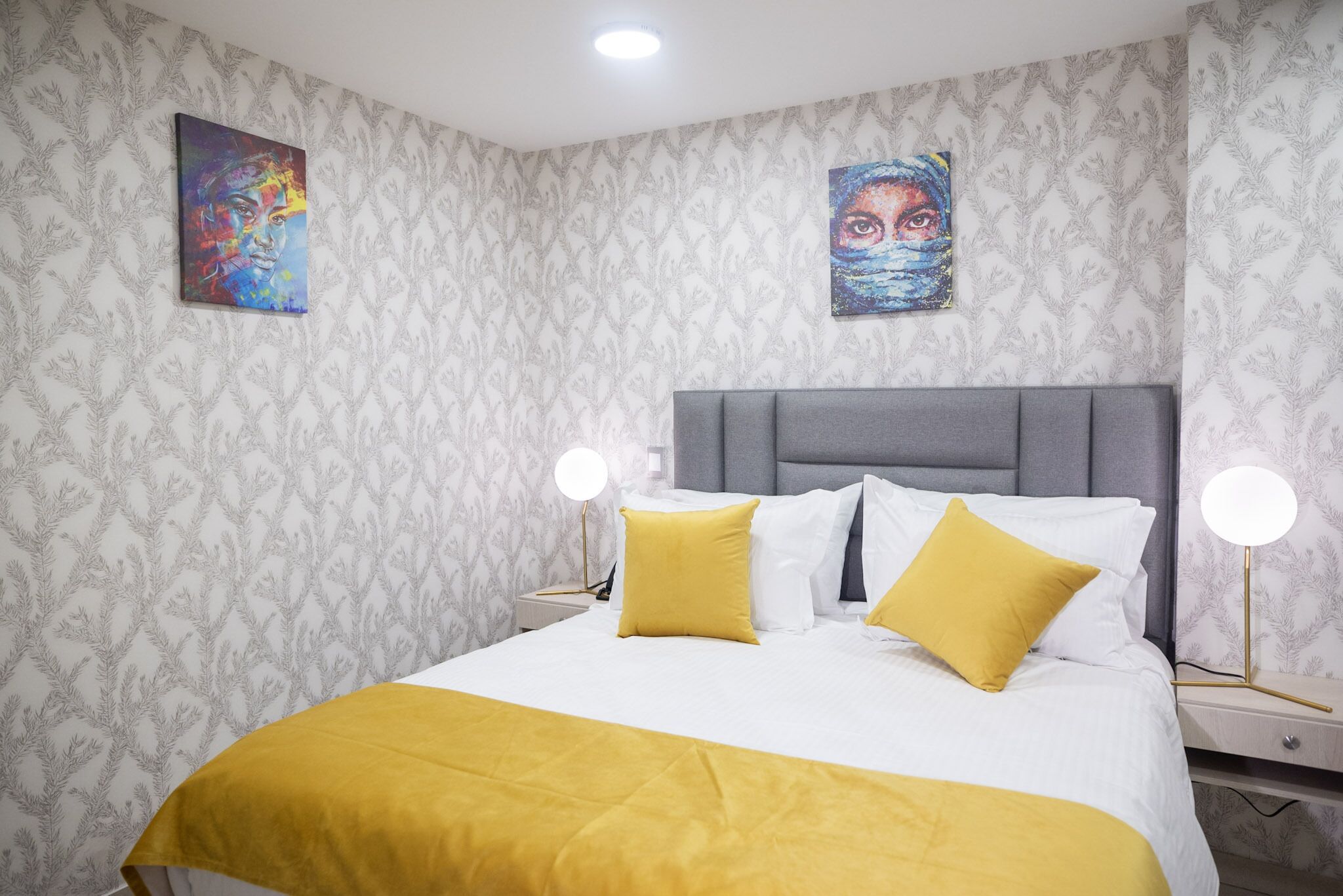 Een opgemaakt bed met een gele sprei en kussens, een grijs hoofdbord en twee bedlampen.