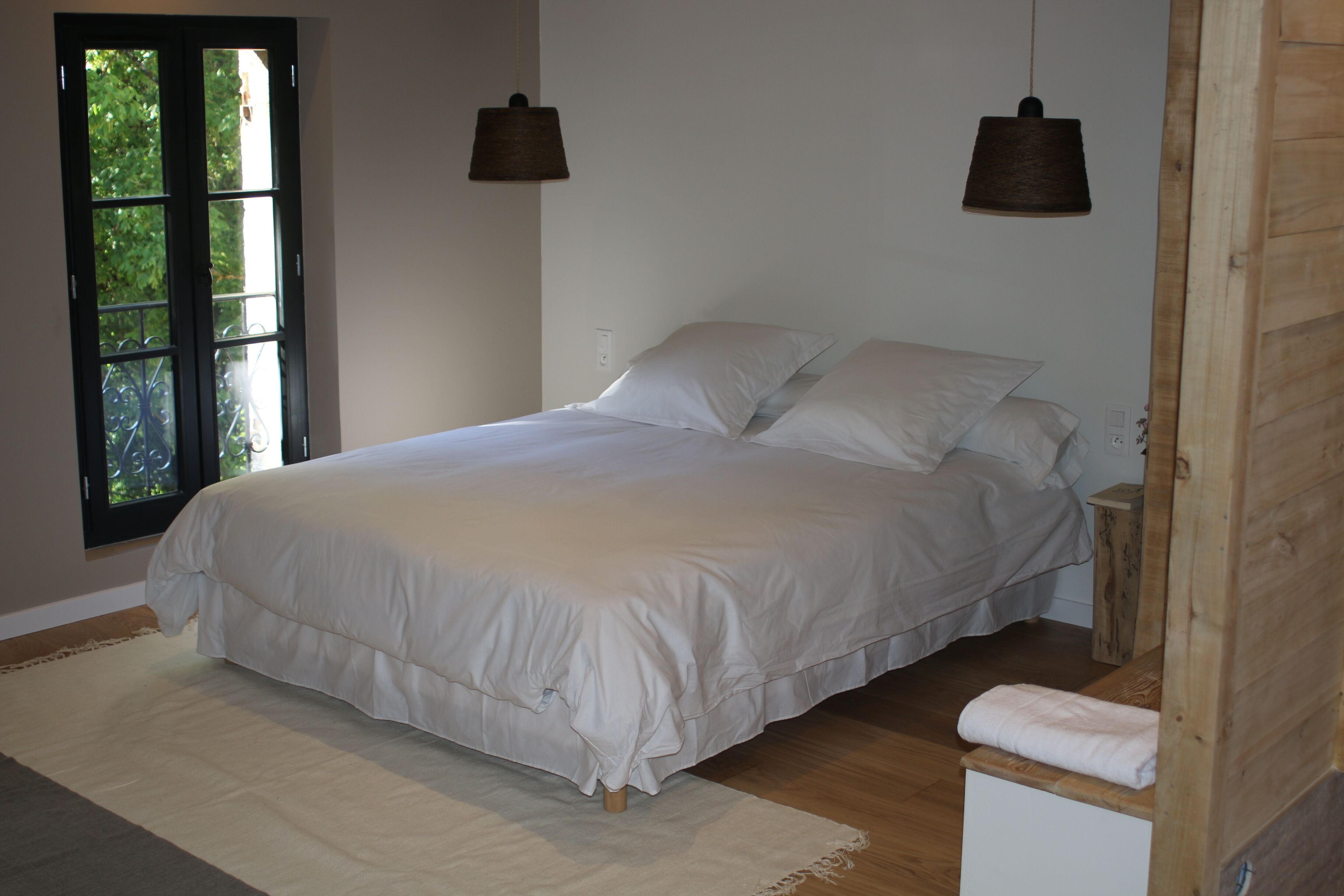 A bedroom with a large bed, two bedside tables, a wooden bench, and a window with a view of greenery.