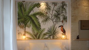 A bedroom with a bed, white bedding, a tropical mural, and a shelf with two wall-mounted lamps. - Malis Castéra Le petit château (Saint-Jean-de-Blaignac)