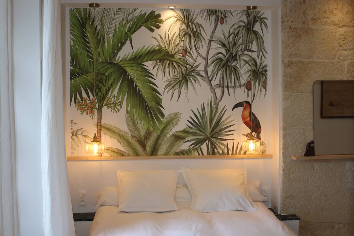 Un dormitorio con cama, ropa de cama blanca, un mural tropical y una estantería con dos lámparas de pared.