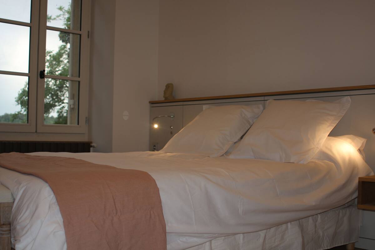Una cama bien hecha con sábanas blancas y una manta rosa, una mesita de noche con una lámpara, y una ventana con vista a árboles.