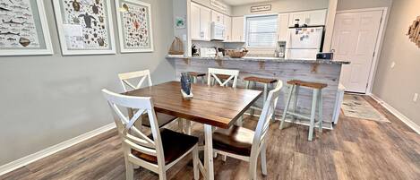 A dining area with a wooden table and white chairs, a kitchen with white cabinets, and framed artwork on the walls.