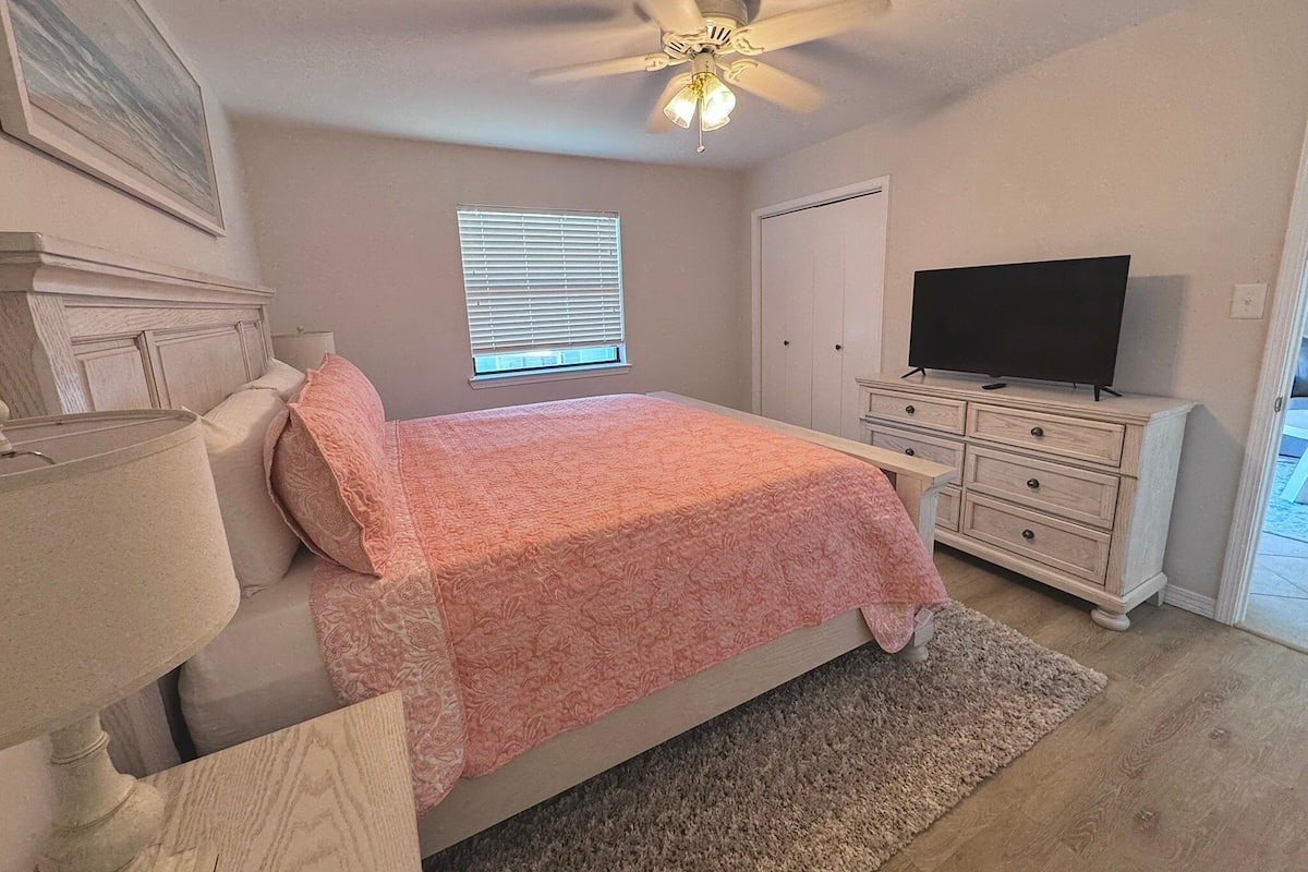 A bedroom with a bed, a ceiling fan, a TV, a dresser, and a window.