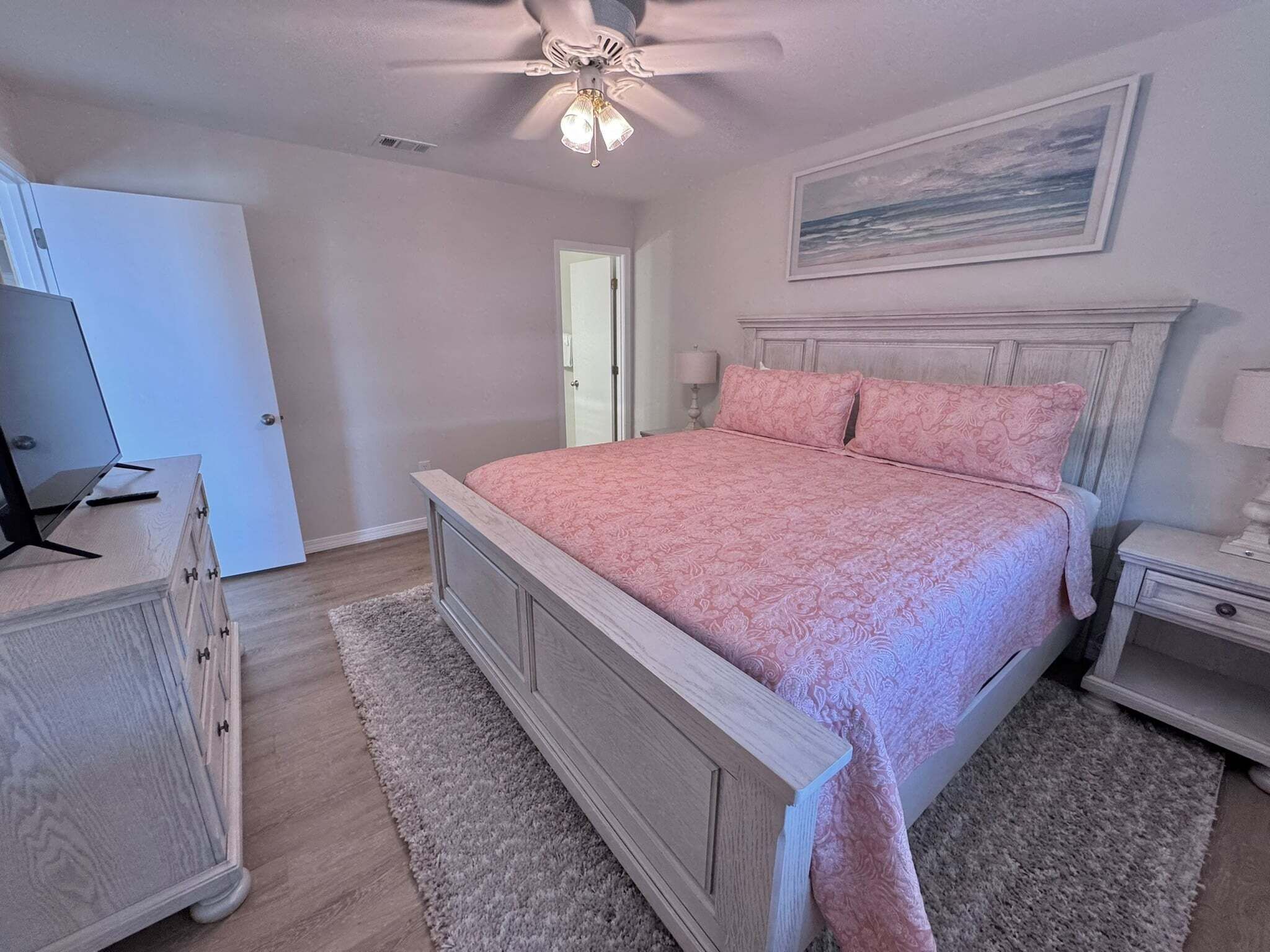 A bedroom with a bed, a nightstand, a dresser, a ceiling fan, a door, and a painting on the wall.