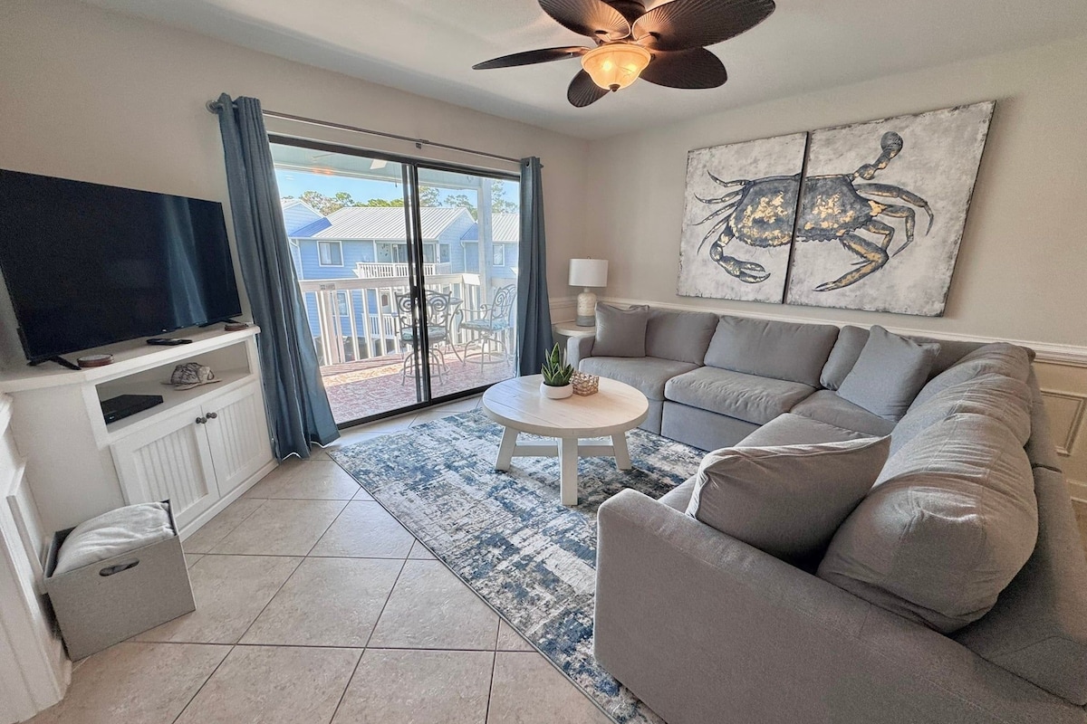 A living room with a sofa, a TV, a ceiling fan, a painting, and a view through a glass door.