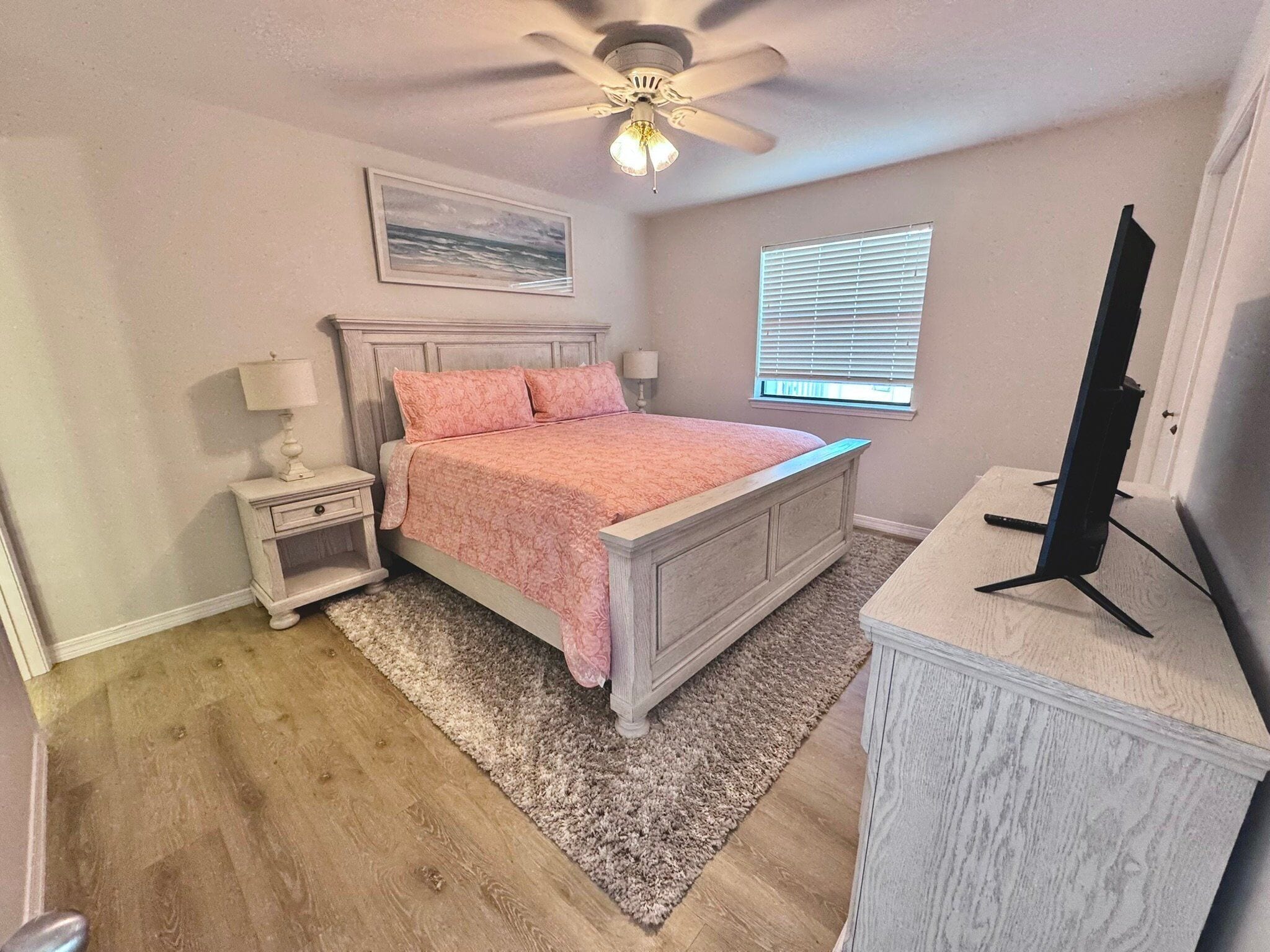 A bedroom with a bed, nightstand, lamp, ceiling fan, window, and a TV stand.
