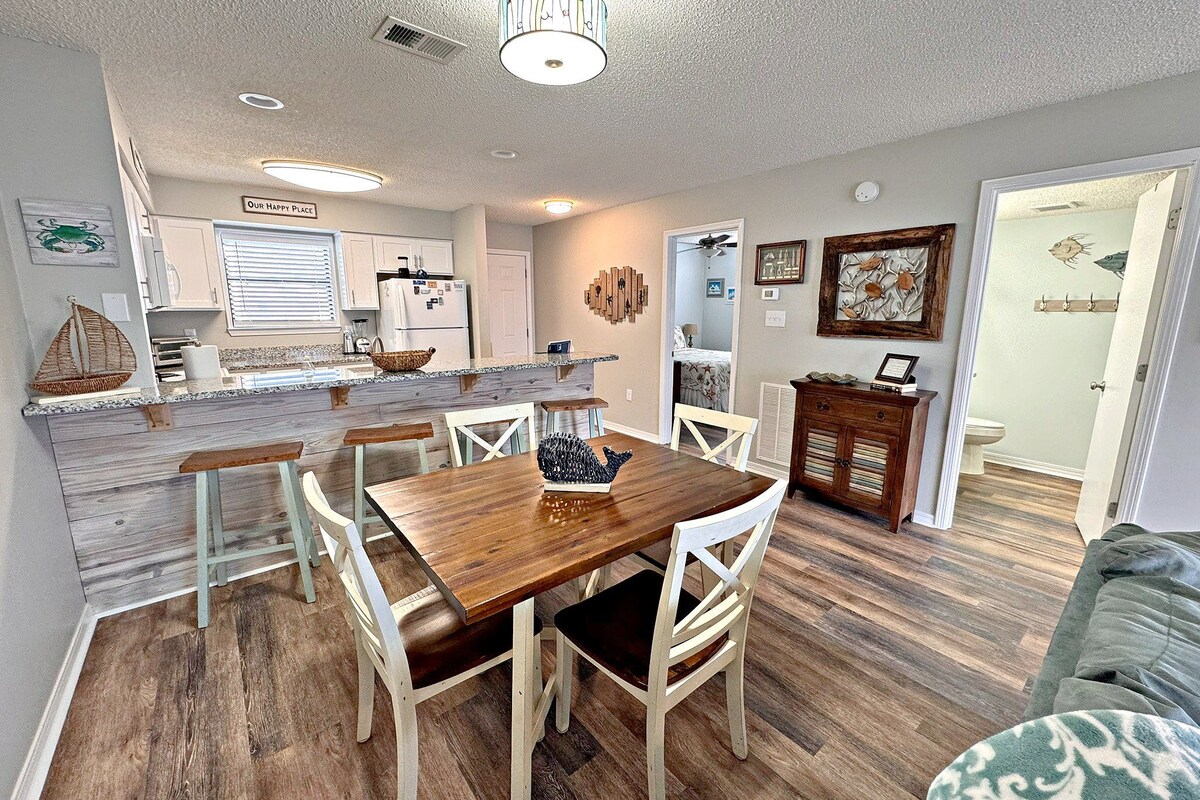 A dining area with a wooden table and white chairs, a kitchen with white cabinets, and a living room with a couch.
