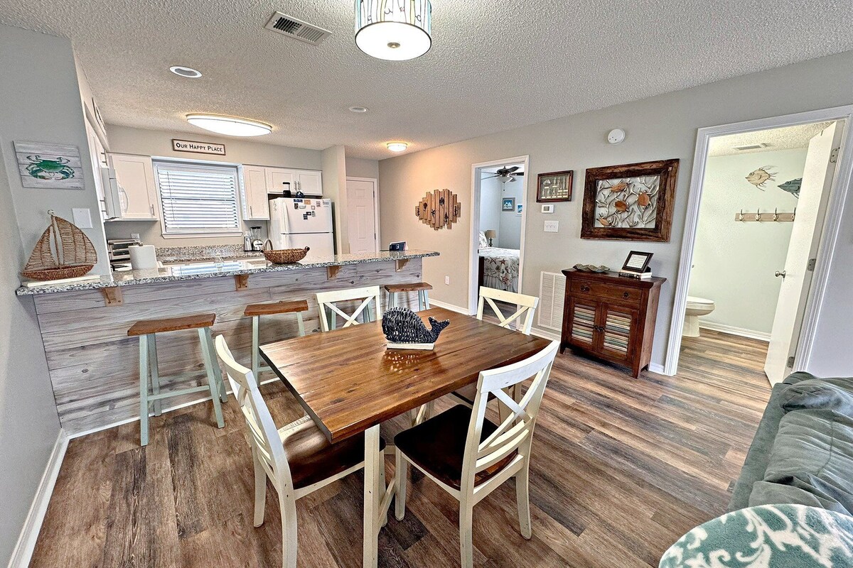 A dining area with a wooden table and white chairs, a kitchen with white cabinets, and a living room with a couch.