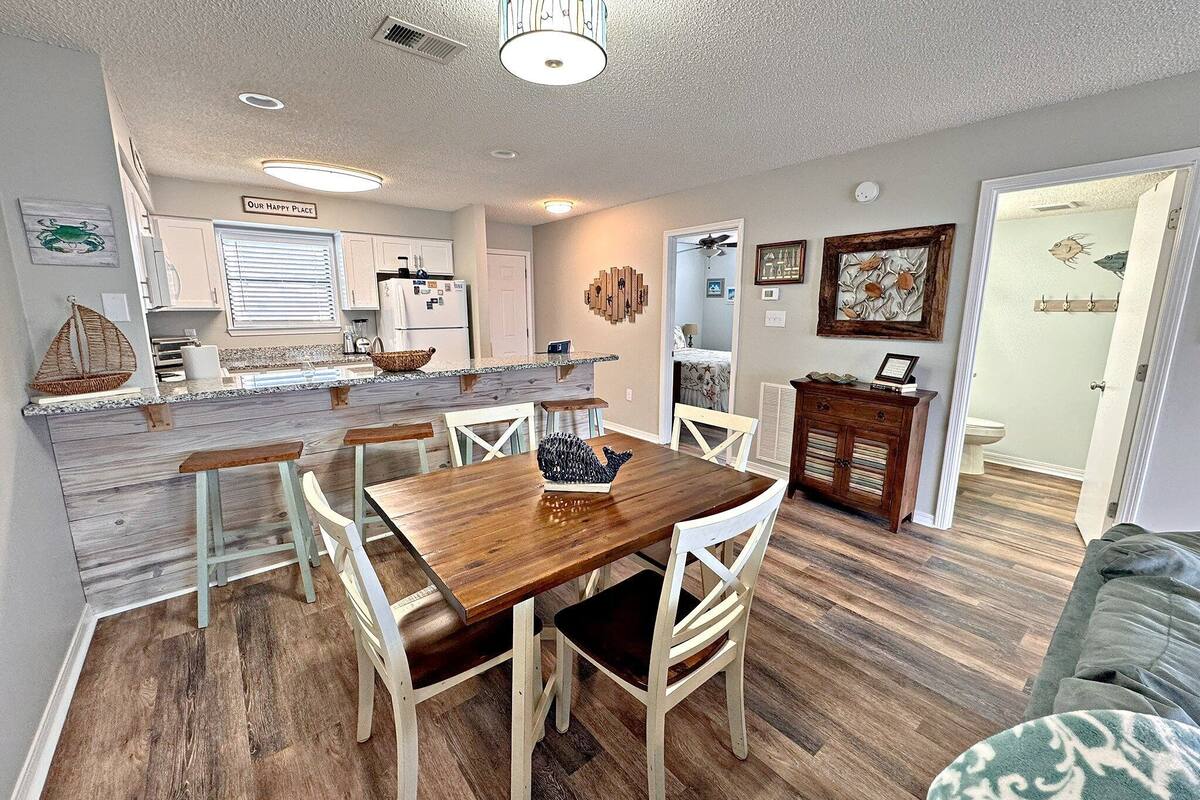 A dining area with a wooden table and white chairs, a kitchen with white cabinets, and a living room with a couch.