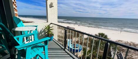 A balcony with turquoise chairs and a table, overlooking a beach with a lifeguard tower and a clear blue sky.