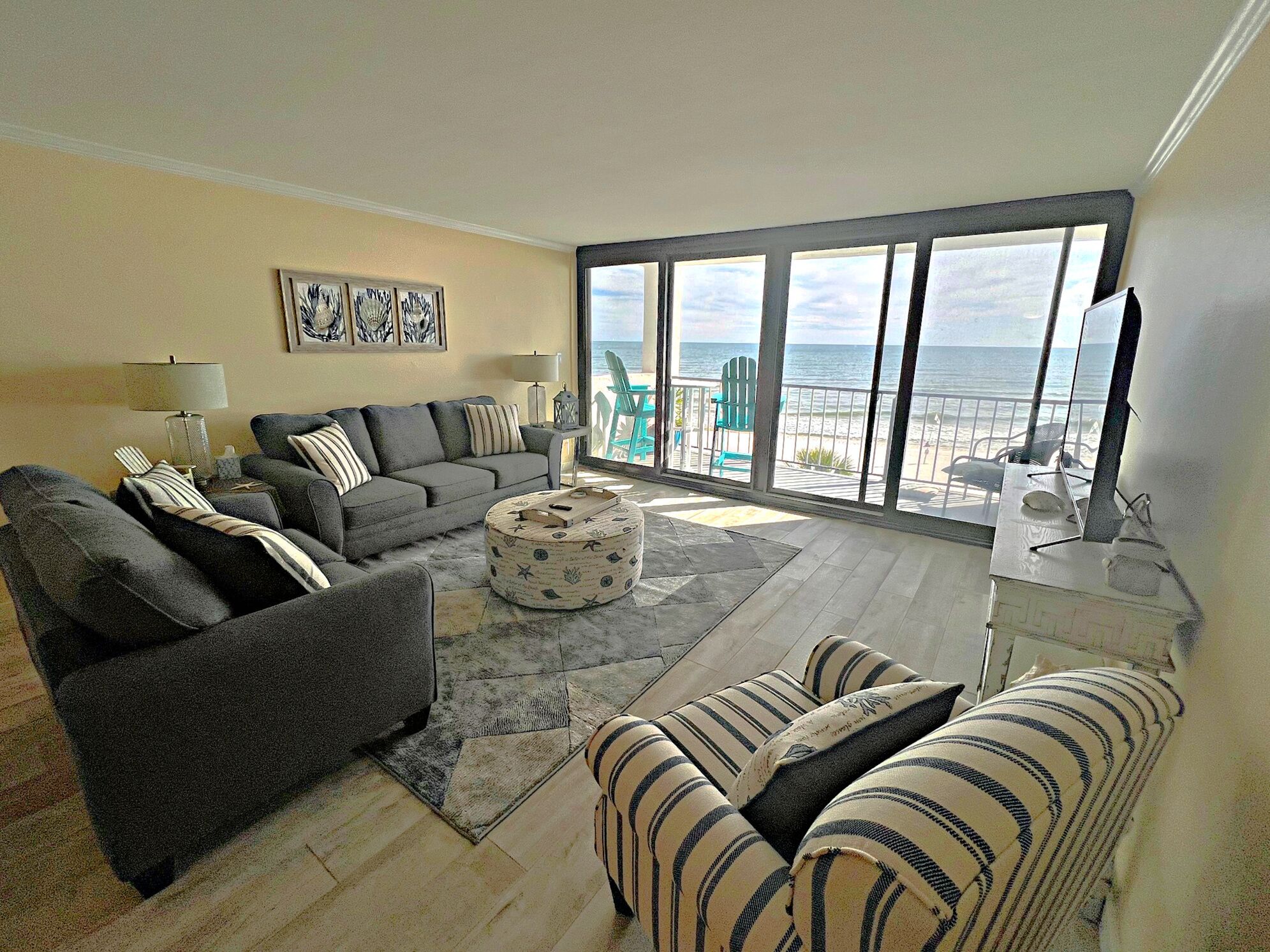 Un salon avec un canapé gris, un pouf à rayures et une vue sur la plage, visible à travers de grandes fenêtres.