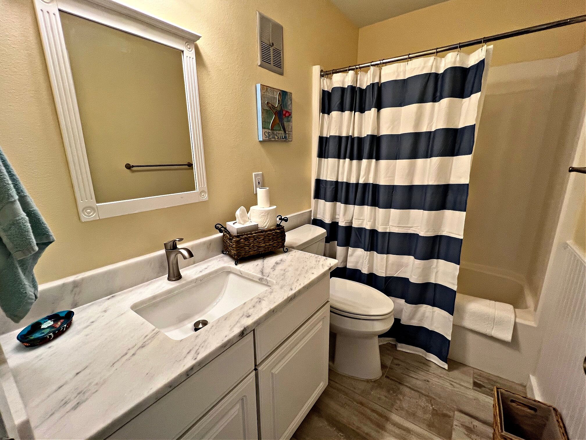 A bathroom with a double sink, a toilet, a shower curtain, a towel rack, and a small painting on the wall.