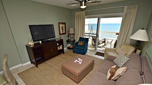 A living room with a television, sofa, ottoman, and a view of the ocean through a balcony. - Caribbean (Gulf Shores)