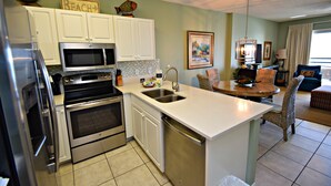 A kitchen with white cabinets, a microwave, an oven, and a dishwasher. There is a dining area with wicker chairs and a table. - Caribbean (Gulf Shores)