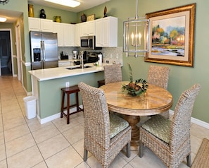 A kitchen with white cabinets, a stainless steel refrigerator, a microwave, and a coffee maker. There is a round dining table with wicker chairs and a centerpiece. - Caribbean (Gulf Shores)