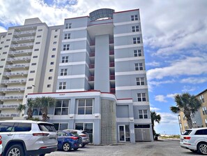 A multi-story building with a central cylindrical tower, multiple balconies, and a parking area with several cars. - Caribbean (Gulf Shores)
