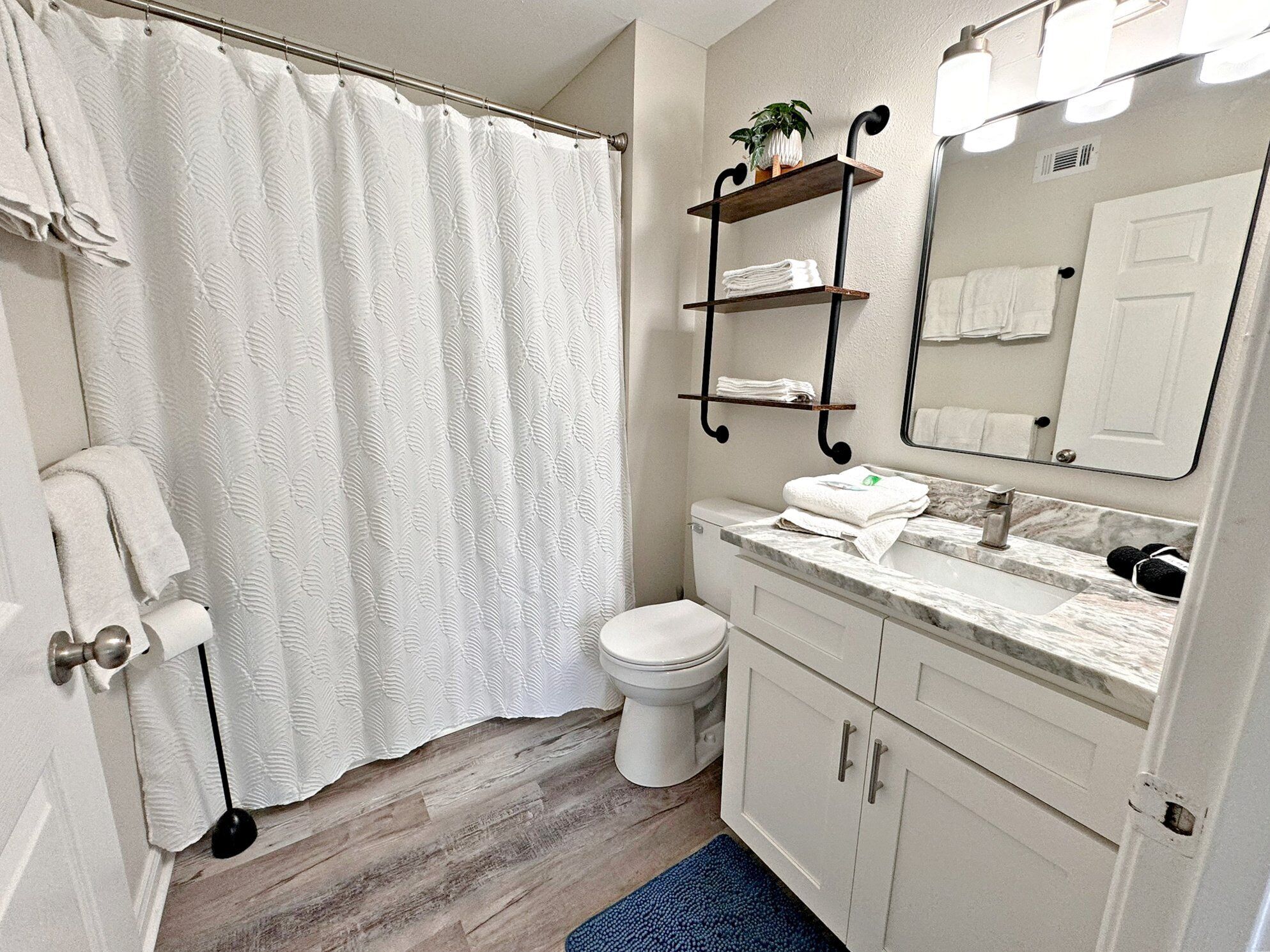 A bathroom with a shower curtain, toilet, sink, mirror, towel rack, and a blue rug.