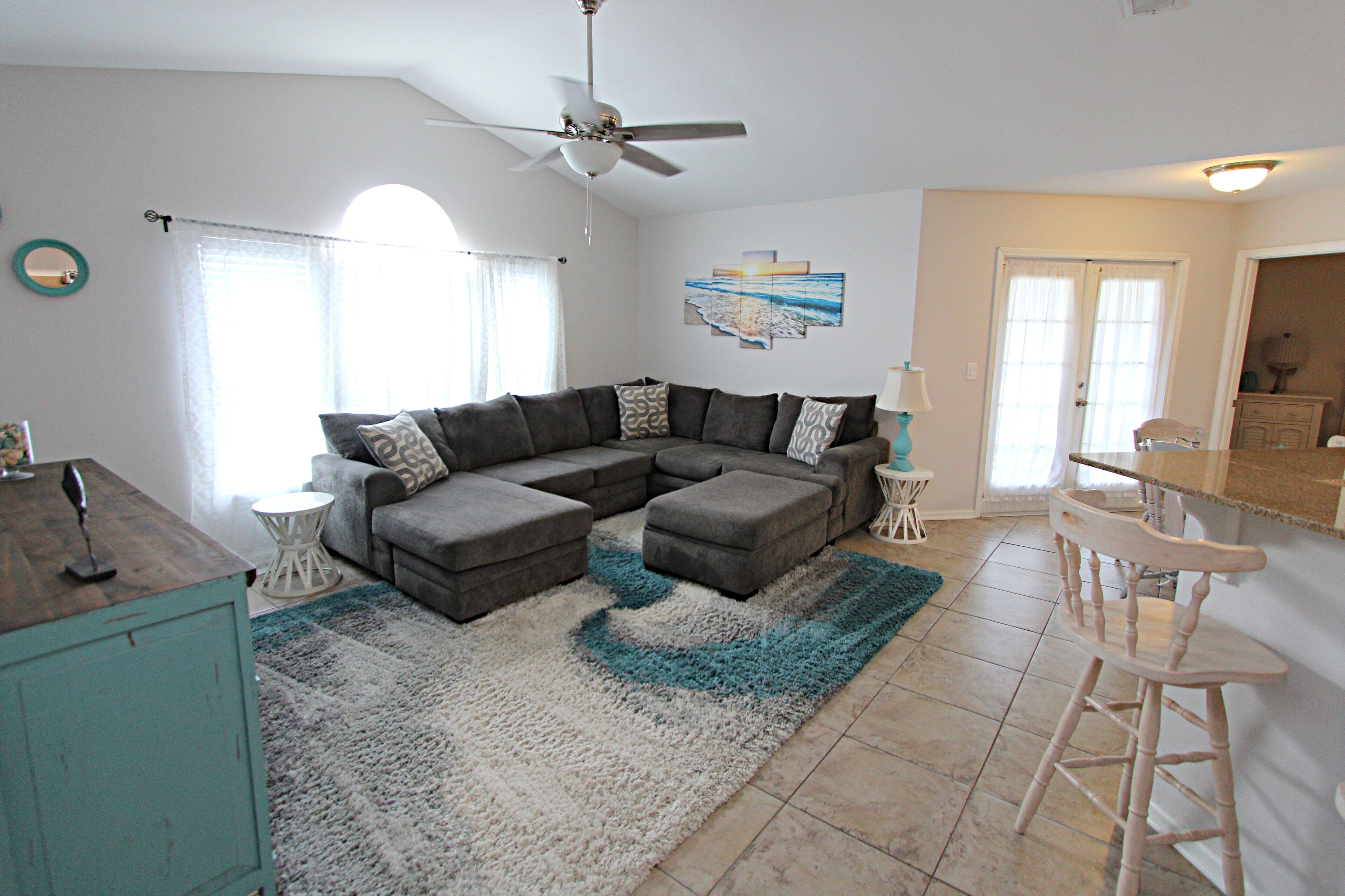 A living room with a sectional sofa, a ceiling fan, and a kitchen area with a bar stool.